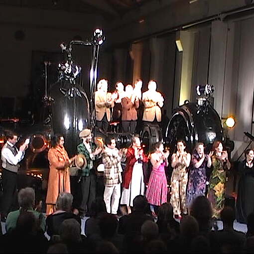 Schauspielergruppe angeleuchtet auf Bühne, die um eine der großen Pumpen angelegt ist